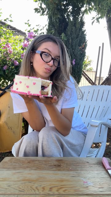 Une part de gâteau individuel coupée en deux, tenue à bout de bras dans un jardin : on voit la crème, la génoise et la garniture à l'intérieur