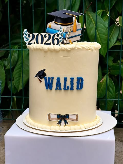Gâteau de remise de diplôme réalisé par Eterna Event : toque, pile de livres et parchemin modelés à la main, prénom et année en pâte à sucre bleue sur un glaçage crème