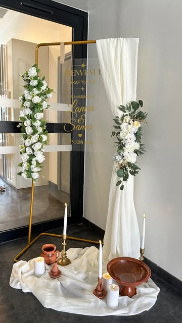 Arche de bienvenue montée par Eterna Event : structure dorée, roses blanches et eucalyptus, drapé ivoire, miroir gravé aux prénoms des mariés, bougies et poterie posées sur un drapé blanc