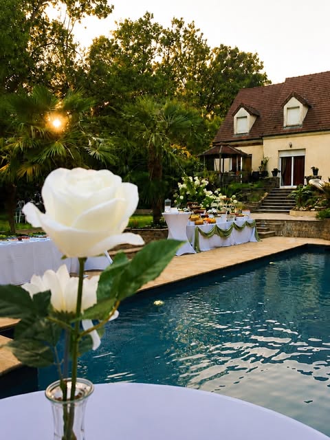 Réception en extérieur montée par Eterna Event au bord d'une piscine à la tombée du jour : deux buffets drapés de vert, roses blanches au premier plan, maison éclairée en fond