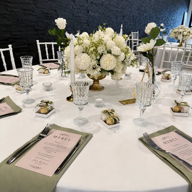 Table de dîner dressée par Eterna Event : nappage blanc, serviettes vert sauge, verres taillés, centre floral blanc dans une coupe dorée, bougies, menus et boîtes de dragées sur chaque assiette